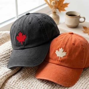 Può includere: Due cappellini da baseball in tessuto di cotone. Un cappellino è grigio antracite con una foglia d'acero rossa ricamata sul pannello frontale. Il secondo cappellino è arancione bruciato con una foglia d'acero color crema ricamata sul pannello frontale. Entrambi i cappellini presentano una visiera curva e una struttura a sei pannelli con un bottone sulla parte superiore.