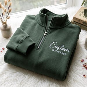 May include: A dark forest green quarter-zip sweatshirt featuring a silver metal zip and ribbed cuffs. The chest area displays white embroidered text that reads Custom Text or Logo in a combination of script and sans-serif fonts.
