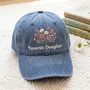 K&ouml;nnte beinhalten: Blaue Jeans-Baseballkappe mit dem gestickten Text "Favorite Daughter" und einem Blumendesign in Rosa, Wei&szlig; und Pfirsich. Die Kappe hat einen gebogenen Schirm und einen Knopf oben.