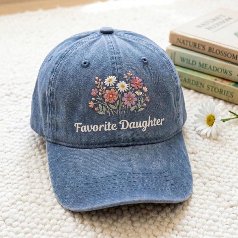 K&ouml;nnte beinhalten: Blaue Jeans-Baseballkappe mit dem gestickten Text "Favorite Daughter" und einem Blumendesign in Rosa, Wei&szlig; und Pfirsich. Die Kappe hat einen gebogenen Schirm und einen Knopf oben.