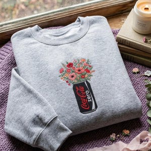 Könnte beinhalten: Ein hellgraues Sweatshirt mit Rundhalsausschnitt und einer Stickerei einer schwarzen Coca-Cola Zero Dose. Die Dose dient als Vase für einen bunten Blumenstrauß aus roten, rosa und orangefarbenen Blüten. Das Motiv ist mittig platziert.