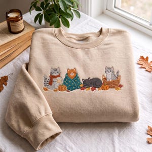 Puede incluir: Una sudadera beige de cuello redondo con un diseño bordado de cuatro gatos en un entorno otoñal. Los gatos aparecen con una taza, envueltos en una manta, durmiendo y sentados en una cesta, rodeados de calabazas naranjas y hojas de otoño.
