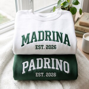 May include: A white sweatshirt with dark green embroidered text reading MADRINA EST. 2026 and a dark green sweatshirt with white embroidered text reading PADRINO EST. 2026. Both garments feature a crew neckline and long sleeves in a folded stack.