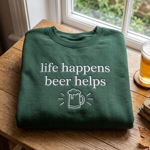 Op de afbeelding: Een donkergroene sweater met ronde hals en wit geborduurde tekst "life happens beer helps" boven een eenvoudige witte lijntekening van een bierpul met schuim, gecentreerd op de borst van het kledingstuk.