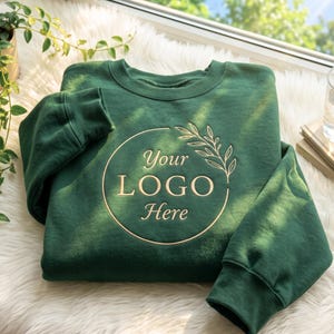 May include: A forest green crewneck sweatshirt featuring a circular gold-toned graphic design on the chest. The design includes a delicate leafy branch element and the text "Your LOGO Here" printed in a clean, serif font centered within the circle.