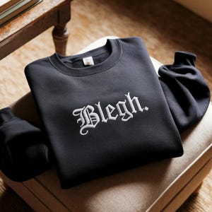 May include: A black long-sleeve crewneck sweatshirt featuring the word "Blegh." embroidered in white gothic-style lettering across the center of the chest. The garment has a ribbed collar, cuffs, and hem, with a visible white interior neck label.