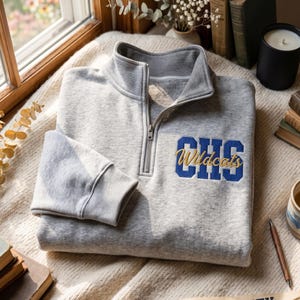 May include: A light gray quarter-zip sweatshirt featuring a high collar and long sleeves with ribbed cuffs. The chest displays embroidered blue block letters reading CHS with the word Wildcats in gold cursive script layered over the letters.