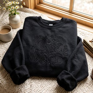 May include: A black crewneck sweatshirt featuring a tonal, embossed-style graphic of four cats in various poses surrounded by botanical elements, stars, and a butterfly. The design is centred on the chest, creating a subtle, textured appearance.