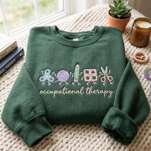 May include: A dark green crewneck sweatshirt featuring an embroidered design of five anthropomorphic objects including a fidget spinner, a ball of yarn, a pencil, a shape sorter, and scissors, with the text occupational therapy embroidered below.