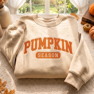 Peut inclure: Un sweat-shirt à col rond beige avec le texte PUMPKIN SEASON brodé en orange sur la poitrine. Le vêtement possède des manches longues avec des poignets côtelés et un col côtelé, fabriqué dans un tissu de couleur unie à la texture lisse.