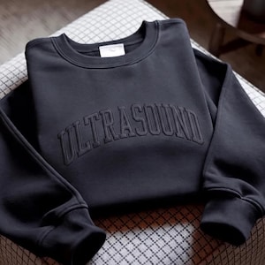 Peut inclure: Un sweat-shirt à col rond gris anthracite avec le mot ULTRASOUND brodé en fil foncé assorti sur la poitrine dans une police universitaire arquée et audacieuse. Le vêtement possède des manches longues avec des poignets et un col côtelés.