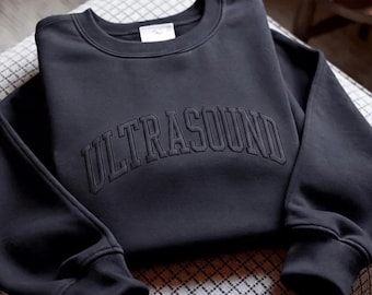 May include: A charcoal gray crewneck sweatshirt featuring the word ULTRASOUND embroidered in a matching dark thread across the chest in a bold, arched collegiate-style font. The garment has long sleeves with ribbed cuffs and a ribbed neckline.
