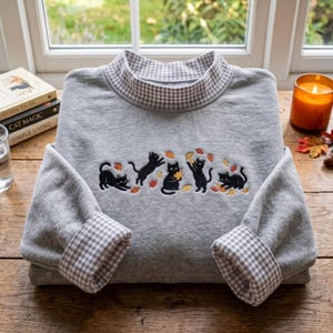 Peut inclure: Un sweat-shirt gris clair doté d'un col montant à motif vichy marron et blanc. Les poignets présentent également le même motif vichy. Cinq chats noirs jouant avec des feuilles orange et jaunes sont brodés sur le devant du vêtement.
