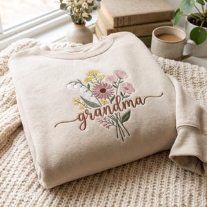 Könnte beinhalten: Ein beigefarbenes Sweatshirt mit Rundhalsausschnitt und einer gestickten Wildblumenbouquet-Applikation in Rosa-, Gelb- und Weißtönen, wobei das Wort "grandma" in brauner Schreibschrift mittig über die Blumen gestickt ist.