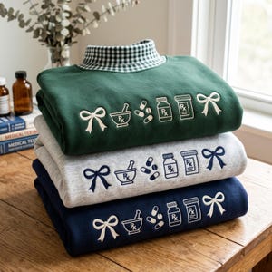 May include: A stack of three sweatshirts in dark green, heather gray, and navy blue. Each features an embroidered design of a bow, a mortar and pestle, pills, and two prescription bottles with Rx labels. The top sweatshirt has a green gingham collar.