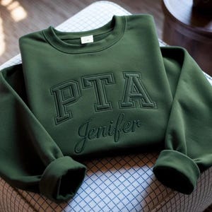 Puede incluir: Una sudadera de cuello redondo en color verde oliva oscuro con el texto PTA bordado en una fuente de bloque en negrita, y el nombre Jenifer bordado debajo en letra cursiva. La prenda tiene mangas largas con puños y cuello acanalados.