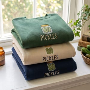 Könnte beinhalten: Ein Stapel aus drei Rundhals-Sweatshirts in Dunkelgrün, Beige und Marineblau. Jedes Sweatshirt zeigt eine gestickte Grafik eines mit grünen Gewürzgurken gefüllten Glases und darunter das Wort PICKLES in einer einfachen, serifenlosen Schrift.
