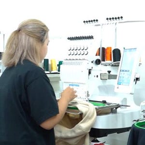 May include: A white industrial embroidery machine with a digital screen and multiple thread spools in various colors. The machine is mounted on a white stand with a lower shelf holding numerous additional spools of thread in a wide array of colors.