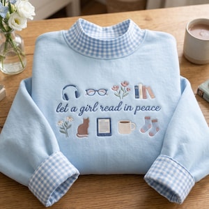 May include: A light blue sweatshirt with a mock neck and cuffs featuring a blue and white gingham pattern. The front is embroidered with the text "let a girl read in peace" surrounded by icons of headphones, glasses, flowers, books, a cat, an e-reader, a mug, and socks.