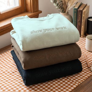 May include: A stack of three crewneck sweatshirts in light green, brown, and black. Each sweatshirt features the text "show your work" embroidered in a matching thread colour across the centre of the chest, with long sleeves and a ribbed neckline.