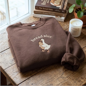 Könnte beinhalten: Ein dunkelbraunes Sweatshirt mit Rundhalsausschnitt und einer Stickerei, die eine weiße Gans mit einem Laib Brot zeigt. Über der Gans ist der Text "bread slut" in cremefarbener Schrift eingestickt. Das Sweatshirt hat lange Ärmel und einen Rippkragen.