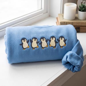 Könnte beinhalten: Ein hellblaues Sweatshirt mit einer Stickerei von fünf Cartoon-Pinguinen in einer Reihe. Jeder Pinguin ist mit einem cremefarbenen Bauch, dunklen Umrissen und orangefarbenen Füßen dargestellt und zeigt verschiedene verspielte Posen auf dem Kleidungsstück.
