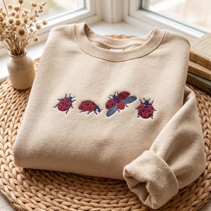 Op de afbeelding: Een beige sweater met ronde hals en een geborduurd ontwerp van vier lieveheersbeestjes op de borst. De lieveheersbeestjes zijn rood met donkerblauwe stippen en details, waarbij er één vliegend is afgebeeld met zichtbare blauwe vleugels.