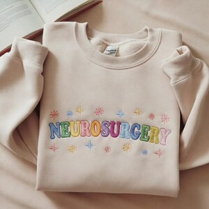 Könnte beinhalten: Ein beigefarbenes Sweatshirt mit Rundhalsausschnitt und dem Wort NEUROSURGERY, das in einer mehrfarbigen, bauchigen Schrift auf der Brust gestickt ist. Das Design wird durch kleine, bunte Sternchen über und unter dem Text ergänzt.