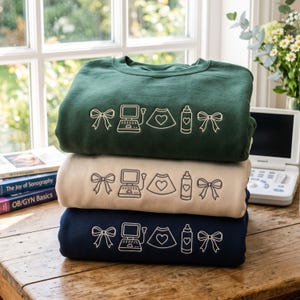 Puede incluir: Una pila de tres sudaderas de cuello redondo en verde oscuro, beige y azul marino. Cada sudadera presenta un diseño idéntico de arte lineal bordado en blanco que consiste en un lazo, un ecógrafo, un escaneo en forma de corazón, un biberón y un lazo.