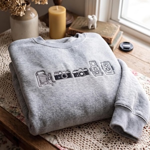 May include: A heather gray crewneck sweatshirt featuring black embroidered line art of five vintage cameras arranged in a horizontal row across the chest. The design includes a folding camera, a rangefinder, an SLR, and two twin-lens cameras.