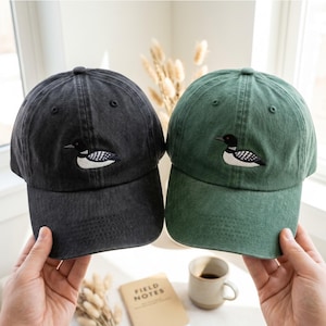 May include: Two baseball caps, one in charcoal gray and one in forest green, each featuring an embroidered loon bird on the front panel. The caps have a washed, vintage-style fabric texture, a curved brim, and stitched eyelets on the crown.