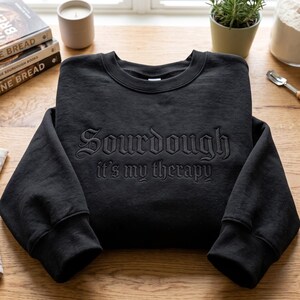 May include: A black long-sleeve crewneck sweatshirt featuring the embroidered text "Sourdough it's my therapy" in a matching black thread across the chest. The garment has a ribbed collar, cuffs, and hem, showcasing a monochromatic design.