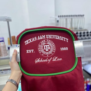 May include: A maroon sweatshirt secured in a green embroidery hoop featuring white embroidered text that reads Texas A&M University, Est. 1989, and School of Law, centered around a circular Texas A&M University logo with the letters ATM.