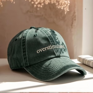 Pode incluir: Um boné de beisebol verde-floresta escuro com um acabamento desbotado de estilo vintage. O painel frontal apresenta a palavra "overstimulated." bordada numa fonte branca sem serifa. O boné tem uma aba curva e uma construção de seis painéis.