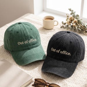 Op de afbeelding: Twee baseballpetten, één in gedempt groen en één in antracietzwart, beide met een constructie van zes panelen, een gebogen klep en een verstelbare band. Elke pet heeft de tekst "Out of office." geborduurd in een lichtbeige kleur.