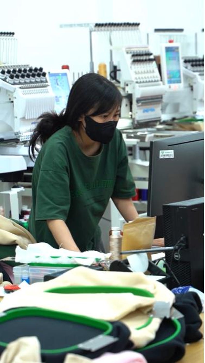 May include: A person wearing a dark green t-shirt and a black face mask, working at a computer workstation in an embroidery facility. The workspace includes industrial embroidery machines, spools of thread, and green embroidery hoops.