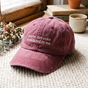 May include: A maroon baseball cap with a distressed finish featuring the embroidered white text "maybe diet coke is addicted to me" on the front panel. The cap has a curved brim, a button on the top, and stitched eyelets for ventilation.