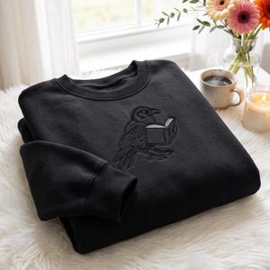 Könnte beinhalten: Ein schwarzer Rundhals-Pullover mit einer detaillierten Stickerei, die einen Raben mit einem offenen Buch zeigt. Das Motiv ist mit schwarzem Garn gestickt, wobei die Buchseiten in Weiß hervorgehoben sind und mittig auf der Brust platziert.