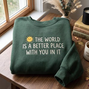 May include: A dark green crewneck sweatshirt featuring an embroidered yellow smiling sun icon and the white text that reads, THE WORLD IS A BETTER PLACE WITH YOU IN IT. The garment has long sleeves and a ribbed collar, cuffs, and hemline.