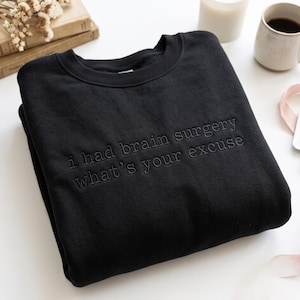 May include: A black crewneck sweatshirt featuring tonal black embroidery on the chest. The text reads, "i had brain surgery what's your excuse" in a simple, sans-serif font. The garment has long sleeves, a ribbed collar, and ribbed cuffs.