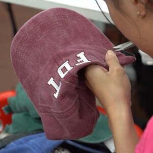May include: A maroon baseball cap with white embroidered letters reading FDT on the front panel. The cap features a curved brim and stitched detailing, held in a position that displays the front design clearly against a blurred background.