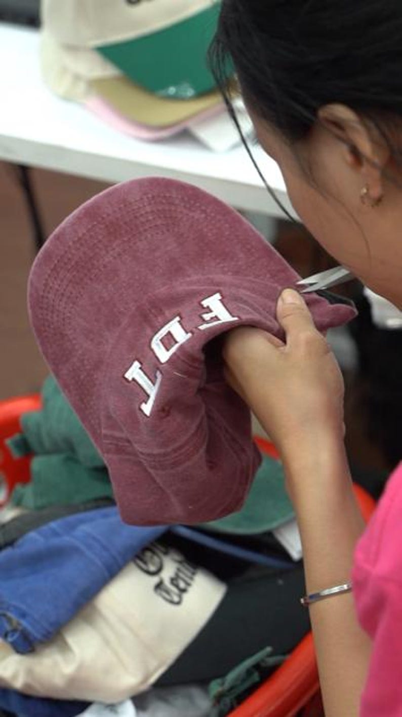 May include: A maroon baseball cap with white embroidered letters reading FDT on the front panel. The cap features a curved brim and stitched detailing, held in a position that displays the front design clearly against a blurred background.