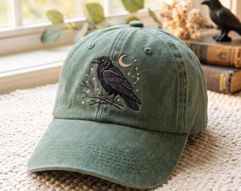 Può includere: Un cappellino da baseball verde salvia con un ricamo raffigurante un corvo nero appollaiato su un ramo, circondato da una luna crescente e piccole stelle. Il cappellino presenta una visiera curva, un bottone superiore e occhielli cuciti.