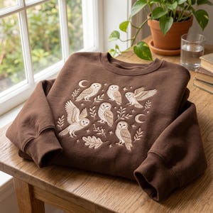 May include: A dark brown crewneck sweatshirt featuring an embroidered design of five owls in various poses, interspersed with small stars, crescent moons, and leafy branches. The embroidery is rendered in light cream and tan thread colours.