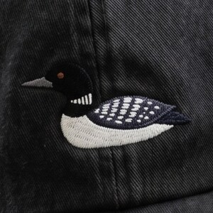 Pu&ograve; includere: Un cappellino da baseball nero con il ricamo di una strolaga maggiore. L'uccello &egrave; raffigurato di profilo con testa nera, una macchia bianca sul collo e corpo scuro con dettagli a macchie bianche. Il tessuto del cappello presenta una trama diagonale.