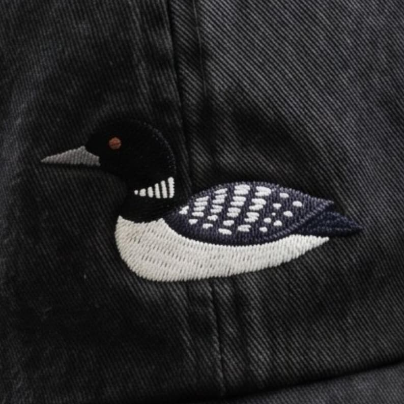 Pu&ograve; includere: Un cappellino da baseball nero con il ricamo di una strolaga maggiore. L'uccello &egrave; raffigurato di profilo con testa nera, una macchia bianca sul collo e corpo scuro con dettagli a macchie bianche. Il tessuto del cappello presenta una trama diagonale.