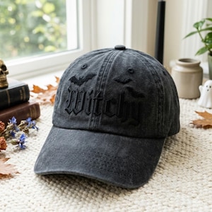 Könnte beinhalten: Eine anthrazitfarbene Baseballkappe mit dem Wort Witchy, das in einer gotischen Schrift auf der Vorderseite eingestickt ist. Das Design enthält kleine gestickte Fledermaussilhouetten über dem Text. Die Kappe hat ein verwaschenes Stofffinish.