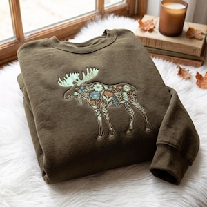 Può includere: Una felpa girocollo verde oliva con una grande silhouette di alce ricamata sul petto. Il design dell'alce è riempito con intricati motivi floreali nelle tonalità dell'azzurro, bianco, arancione e verde salvia, con palchi verde chiaro.