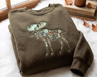 May include: An olive green crewneck sweatshirt featuring a large embroidered moose silhouette on the chest. The moose design is filled with intricate floral patterns in shades of light blue, white, orange, and sage green, with light green antlers.
