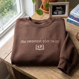 May include: A dark brown crewneck sweatshirt featuring the embroidered text "the sweetest plot twist" in a light beige thread, positioned above a small, matching embroidered icon of an open book with two tiny baby footprints on the pages.